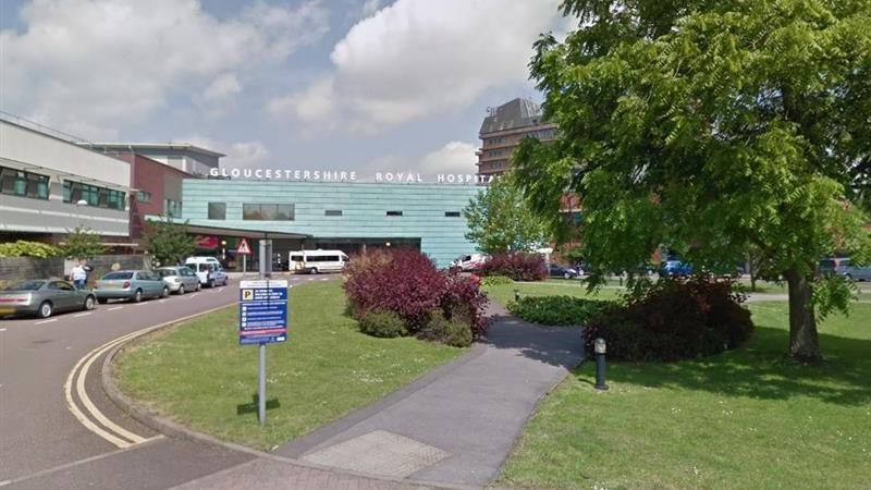 Image of the hospital entrance. The words Gloucestershire Royal Hospital appear in white above the turquoise tiles at the front ofthe building. a blue directional sign is in the foreground.