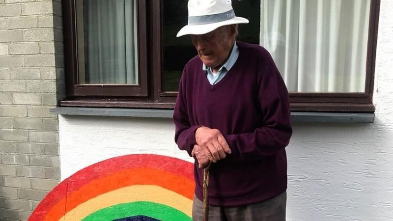 Rhythwyn Evans standing outside a brick wall painted with a rainbow. He is wearing a v neck maroon jumper, a light coloured shire and a white brimmed hat. He is holding a cane in one hand and had his right hand holding his left wrist 