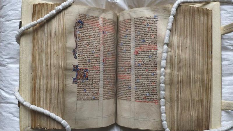 The medieval manuscript is held open so a double-page spread can be seen. There are two decorated capital letters in red and blue and some coloured writing in amongst the need medieval Latin.