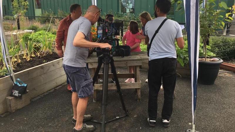 The back of a man holding a camera who is filming pupils and presenter Justin Fletcher