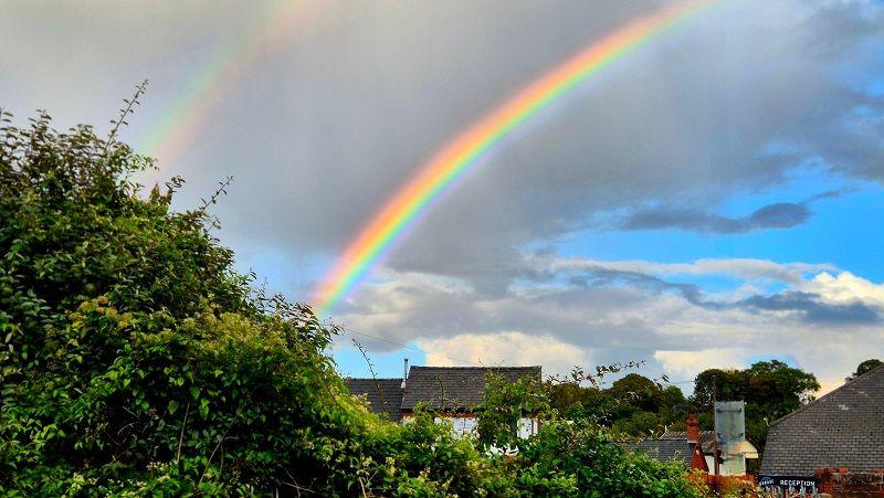 Two rainbows curve through the sky over rooftops in a town, the sky is overcast with grey clouds with some blue popping through