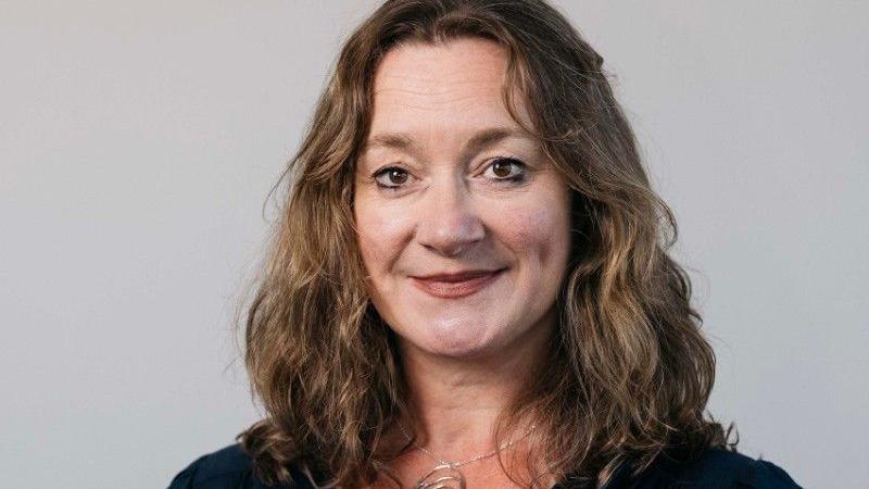Daphne Caine, who has shoulder-length light brown wavy hair. She is wearing a black town and has thin necklace around her neck, against a grey background.