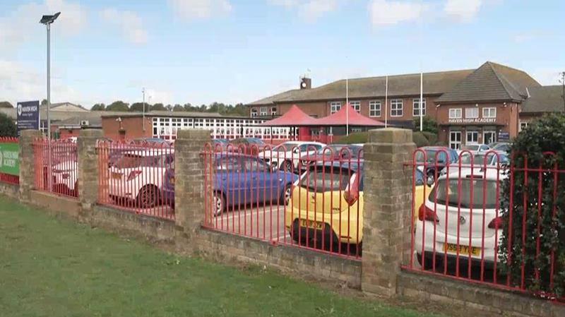 A secondary school comprising a mix of single and two-storey buildings and a large red canopy over the entrance. Cars are parked in a car park. In front of the car park, nearest the camera lens, is the school field.   