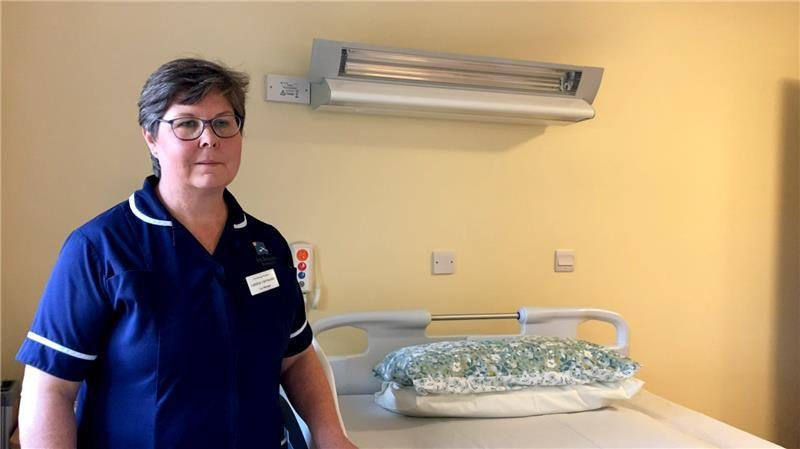 A nurse with short grey hair and glasses in a blue nurse's dress. She in next to a hospital bed.