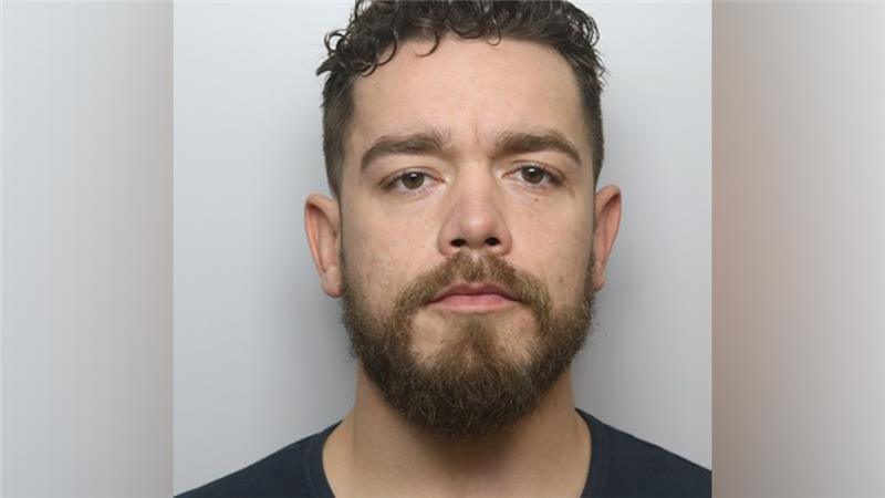 A mugshot of a man with short black hair, a dark beard and a black shirt.