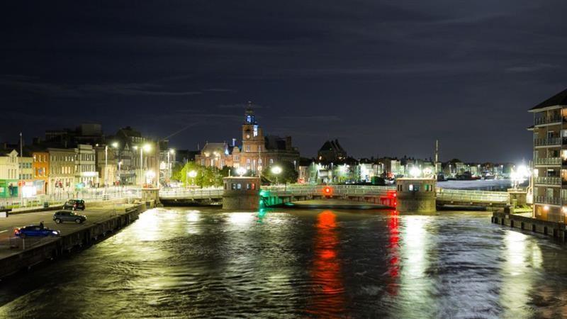 Haven Bridge at night. There are town houses to the left on the banks of the river and some parked cars. There are clouds above.
