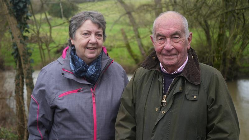 A man and a woman are standing next to each other by the river Waveney. The woman is wearing a purple raincoat and the man is wearing 