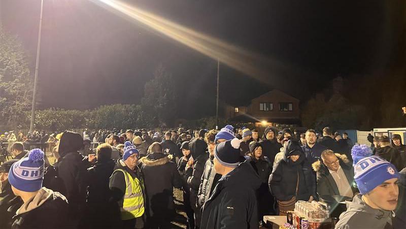 A large crowd of football fans, some in blue Daisy Hill woolly hats, wait for kick-off