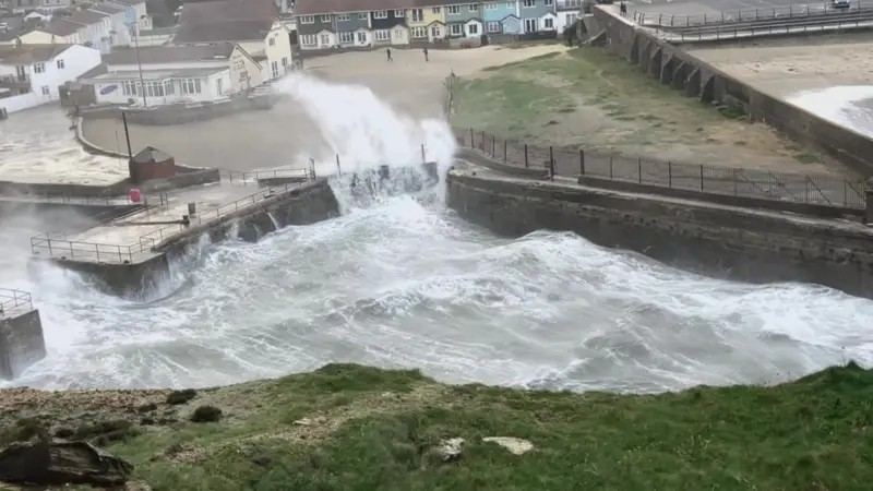 Large waves engulf the wall and railings of a small Cornish harbour. Houses behind and a bank of grass in the foreground.