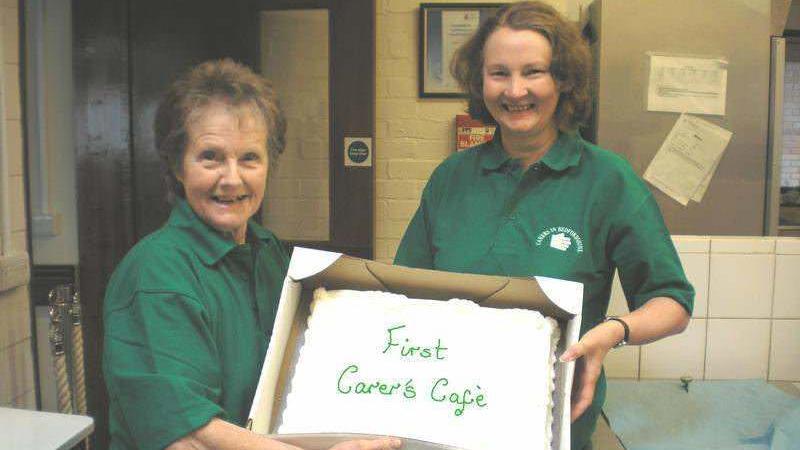 Yvonne Clark stands beside a second woman, they are both wearing green tops which read "Carers in Bedfordshire". They are holding a large white cake, in a box which has "First Carer's Cafe" decorated on it. They are both in a kitchen and looking at the camera and smiling.