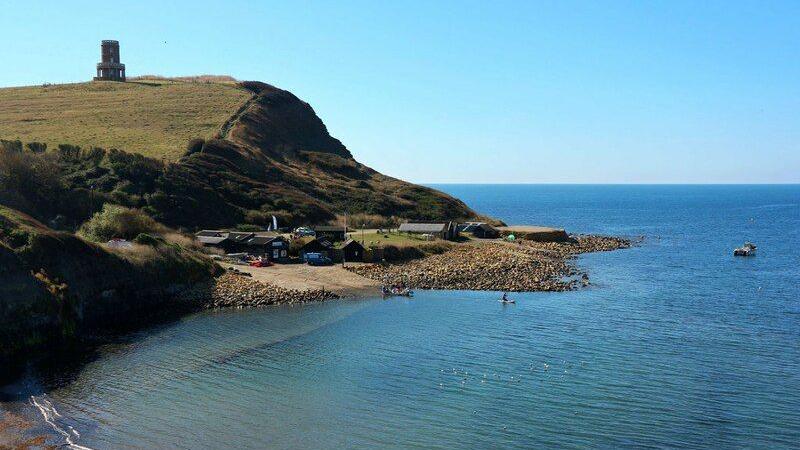 View across Kimmeridge Bay. To the left of the picture is the cliff with Clavell Tower - a round folly - at the top. Beneath is a slipway with some buildings at the top. In the foreground is the shore. The right of the photo is just clear blue sky and vivid blue sea.