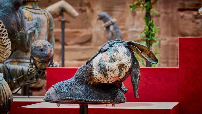 A bust of a human-like figure with rabbit ears and a rabbit face in an outdoor area with a red brick wall behind it and a red display stand directly behind the bust