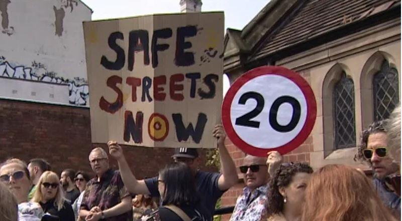 A crowd of about 13 people outside what looks like a church in a street. One man holds up a sign saying Safe Streets Now, another man holds up a 20mph speed limit sign.