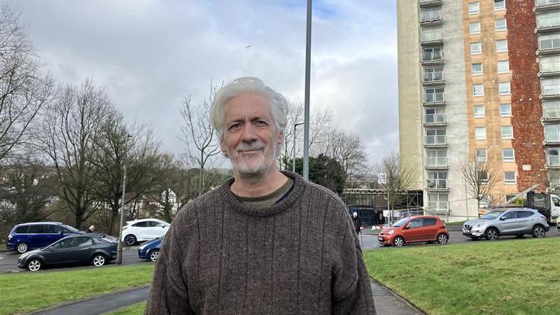 A man wearing a jumper stood in front of a housing block. There are a handful of cars in the background, as well as an area of grass. 