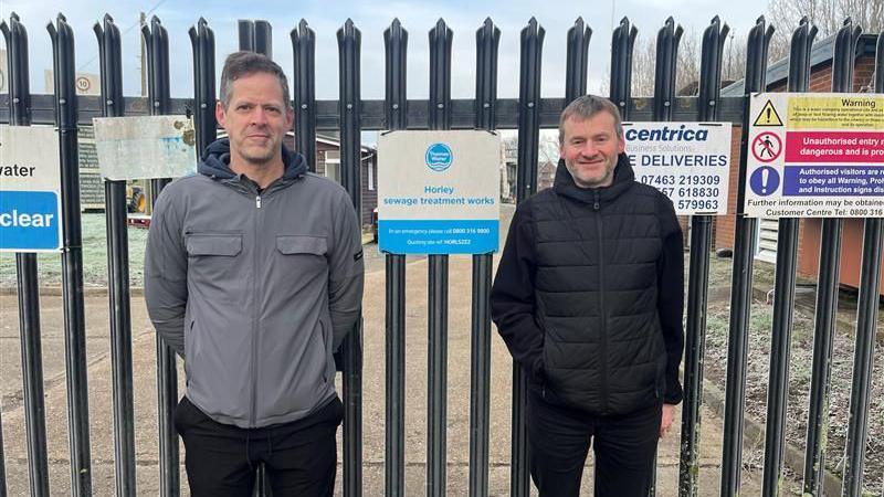 Steve Pelham on the right in a black coat. He has grey hair with a beard and stubble. Local councillor Steve Wotton is on the left in a grey coat, with grey and black hair. They stand in front of the Horley sewage treatment works.