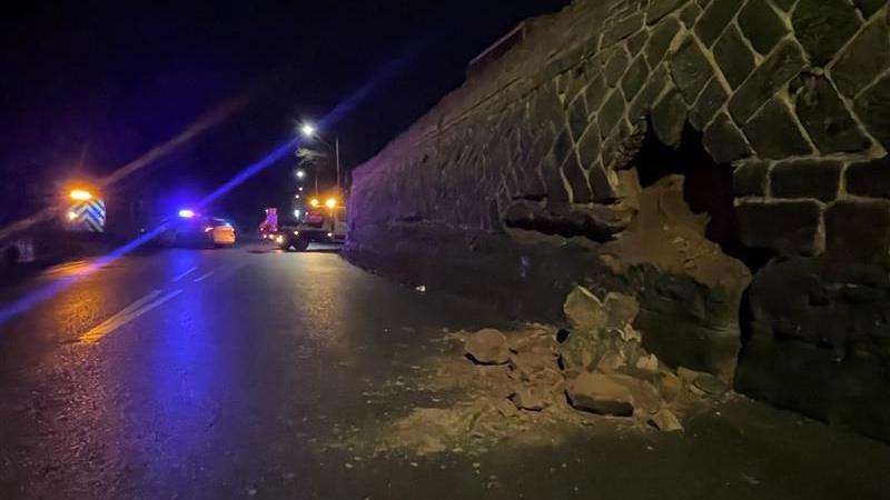 A section of collapsed wall strewn onto the road with emergency services blocking traffic in the distance