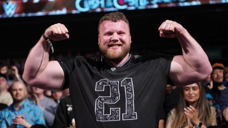 Tom Stoltman wearing a black t-shirt and flexing for the camera.