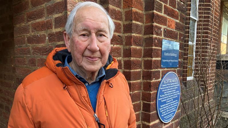 Chris Tong in an orange coat, has short grey hair. Behind him is a red brick wall with a blue plaque on it.