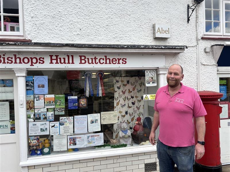 A man in a pink shirt and blue jeans stands outside a white shop with a red sign whose window is full of posters and a model of a chicken and a pig. 