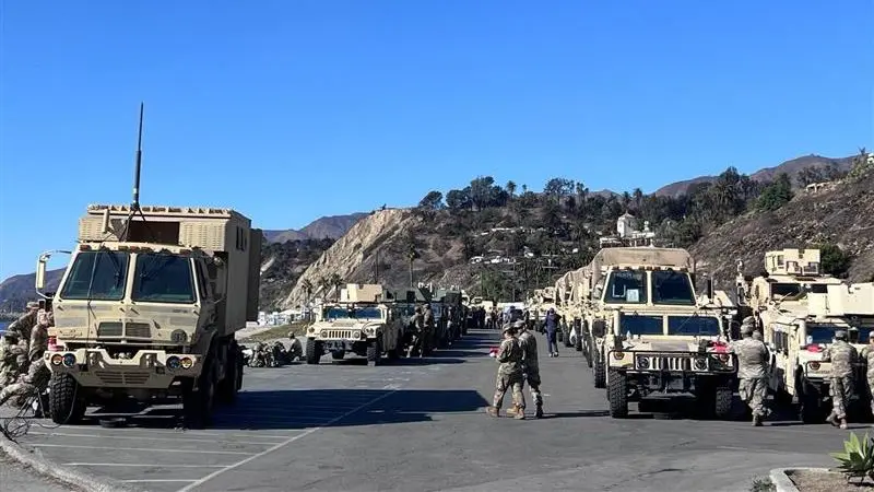 A long line of military vehicles with scattered personnel, in front of hills