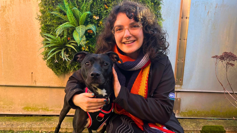 A young woman with curly dark hair, a bright scarf and dark coat, smiles at the camera as she hugs a black dog, which is a type of staffordshire bull terrier cross