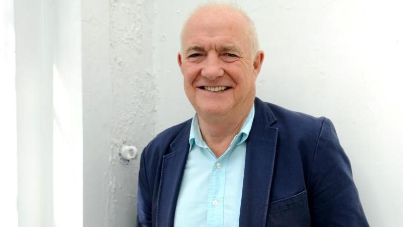 A man with very short blonde hair wearing a light blue shirt and a dark blue blazer stood in front of a white wall.