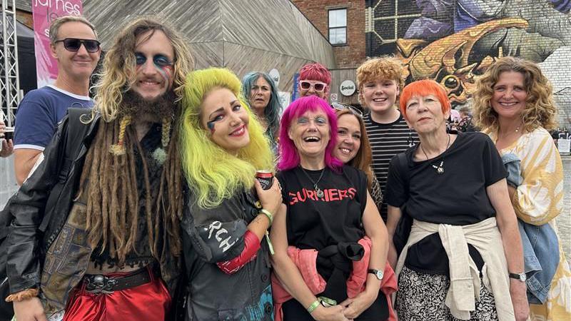 A group of people dressed in colourful outfits with colourful hair. They are standing on a street smiling at the camera. The walls behind them are covered in graffiti. 