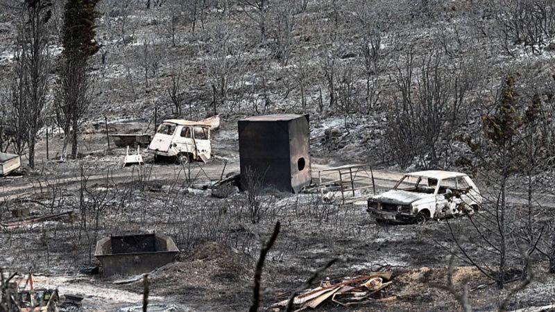 A scene of devastation after a wildfire swept through an area. everything is covered in ash and all vegetation is a black cinder. In the centre of shot, two burnt out cars sit.
