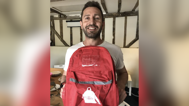 Luke Simon is stood in the middle of the frame, holding a red rucksack. He is smiling at the camera and appears to be in a farmhouse-style room with timber beams on the wall.