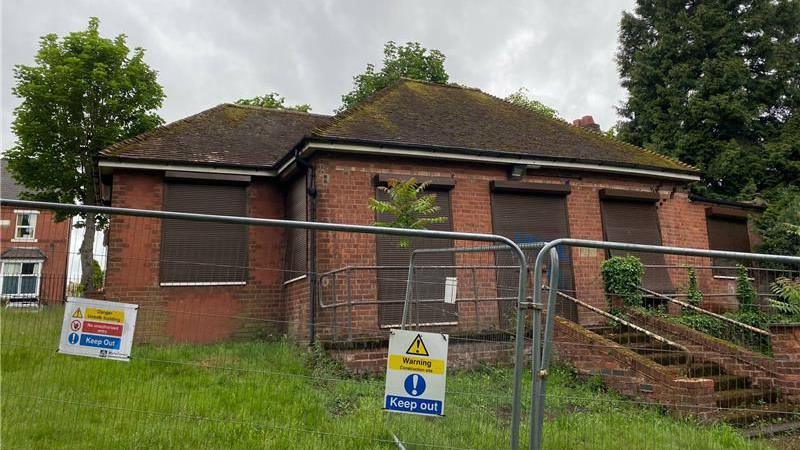 The old pavilion is seen with barriers and keep out signs around it. The windows are boarded up and the building is in disrepair.