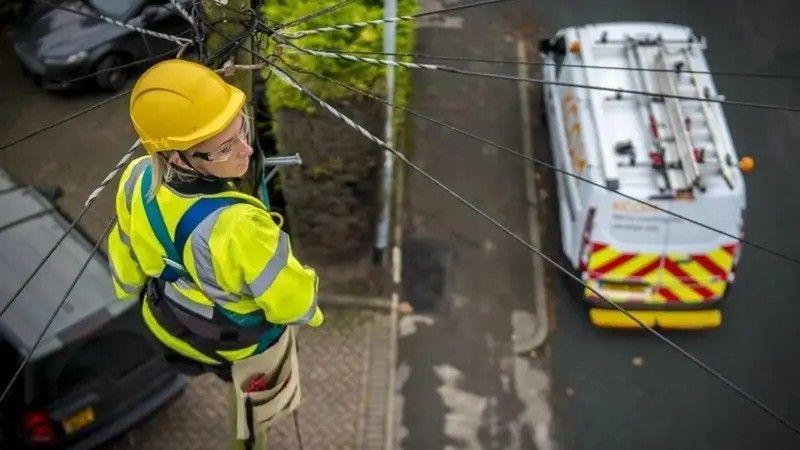 A woman in a hi-vis vest and wearing a helmet and a utility belt up a telegraph pole. There is a van with yellow and red chevrons painted on its back doors parked on the road below.