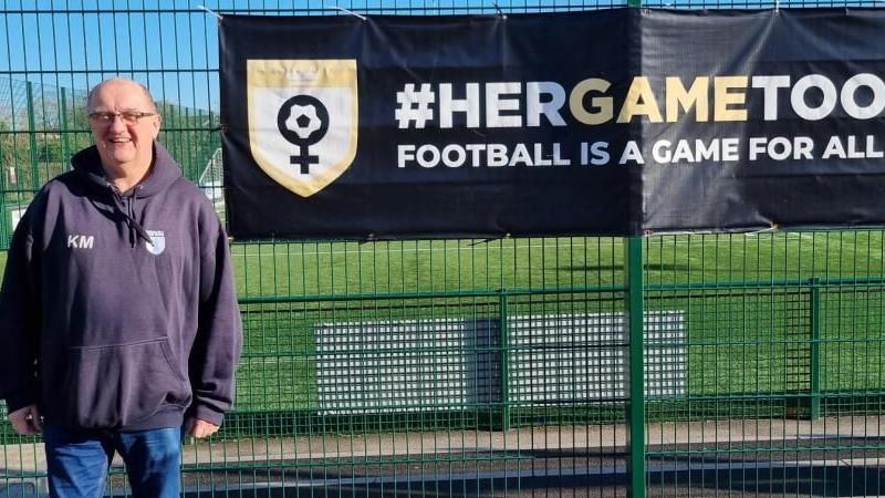 A man wearing jeans and a baggy dark hoodie standing next to a green fence with a black Her Game Too sign written in bold white letters on it. There's a football pitch behind the fence.