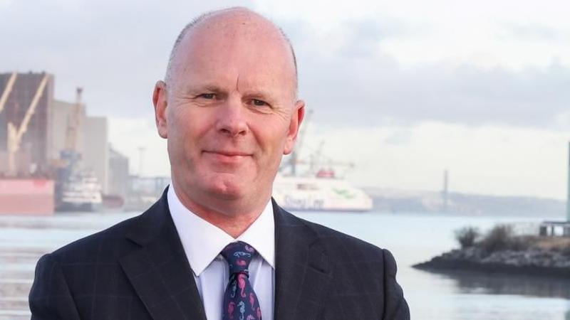 Belfast Harbour Chief Executive Joe O'Neill wearing a black suit, white shirt and a tie with seahorses, standing at Belfast Harbour. A cruise ship and cranes are in the background.