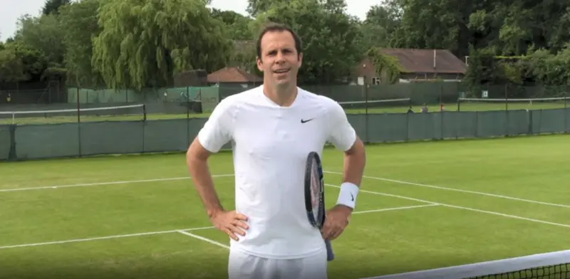Tennis player Greg Rusedski standing on a tennis court, wearing white t shirt and shorts, holding a tennis racket and facing the camera.