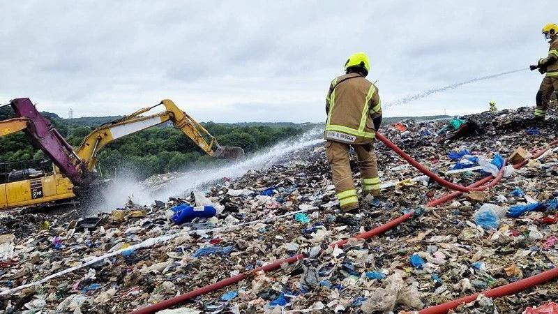 Two firefighters holding fire hoses spraying water at a fire on an area of rubbish at a landfill site.