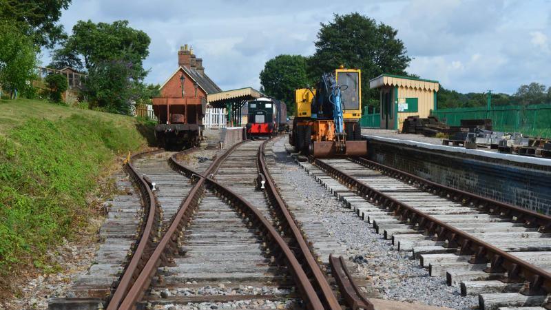 Another view of the restored Shillingstone station. There is a black train sat on the track and a yellow truck on the opposite track working on the site. In the distance is the station property with the green and white awning over the platform. In the foreground of the image is more track and a grass verge to the left.