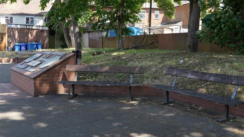 An area of seating in Upper Orwell Street in Ipswich, with trees in the background and paving in the foreground. Sun is shining through leaves in the trees, leaving most of the area under shade