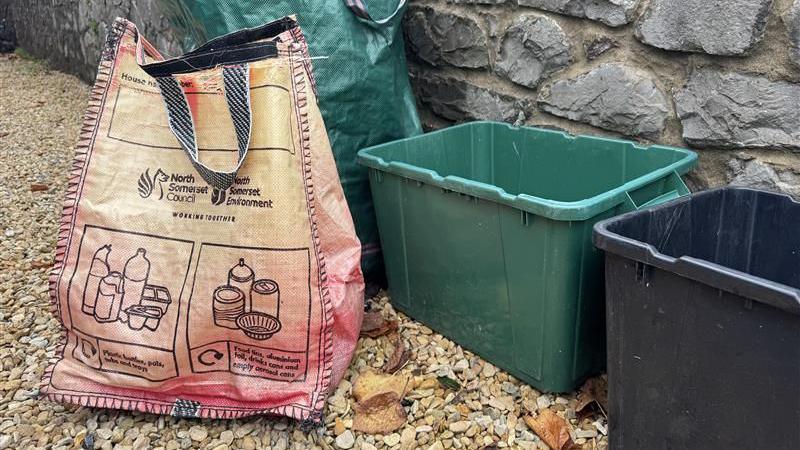 Faded red North Somerset recycling bag, near a green and black recycling box outside on a pebbled street.