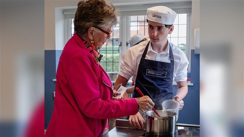 A pupil stands at a hob workstation talking to Prue. He lifts a plastic jug while Prue is stirring something inside a silver saucepan. She wears a dark pink coat. The student wears a white shirt with rolled up sleeves and a white chef's hat.