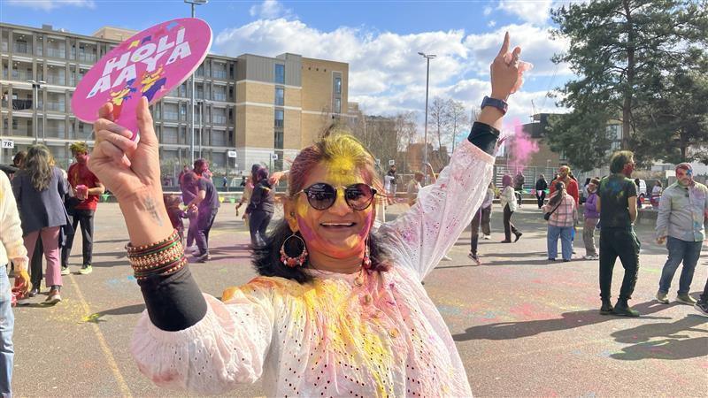 A lady dressed in white and covered in colourful powder wearing sunglasses poses and smiles. There are people covered in paint behind her. 