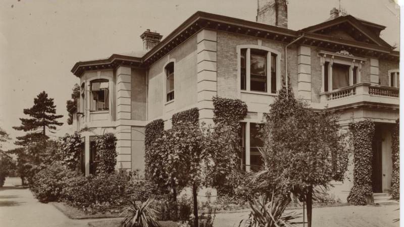 A picture of a large 19th Century country house, partly covered in ivy, with a grand balcony over a doorway, with shrubbery and pathways in view