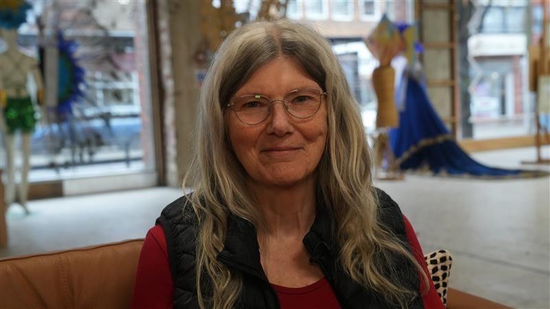 Sue Lawson is smiling at the camera. She has long blonde hair and is wearing a red top and a black jacket. She is sitting in a shop area on a sofa.