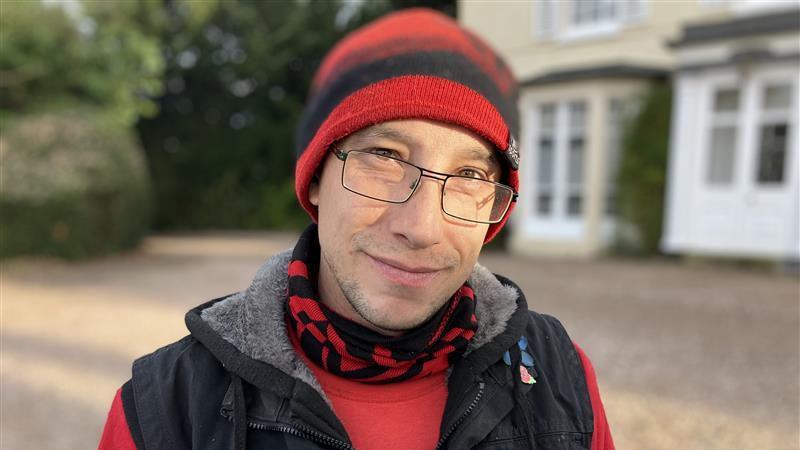 A man standing in front of a house wearing a red and black beanie, glasses, a red and black scarf, a black jacket and a red top.