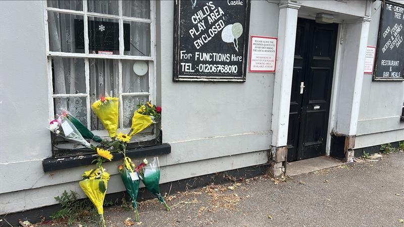 Flowers on the pavement outside the Cherry Tree pub, which is a grey building with a black door.