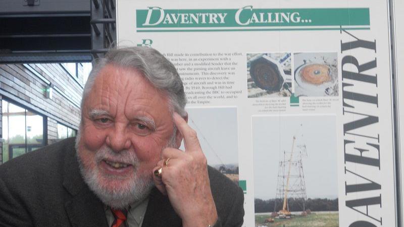 Terry Waite with grey hair and beard wearing a dark jacket and red tie. A finger on his left hand is touching his forehead. There is a gold ring on another finger. He is in front of an information board with "Daventry Calling" at the top and text and photos. There is a window behind him revealing the front of a building.