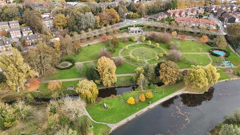 An aerial image of landscaped gardens, mostly laid to lawn with mature trees and pathways. Centre-right is a pathed, circular labyrinth/turf maze  in the grass. To the bottom right of the image is the dark water of the River Wensum. To the top of the image, the park is bordered by a road running left to right, with housing and trees beyond. 