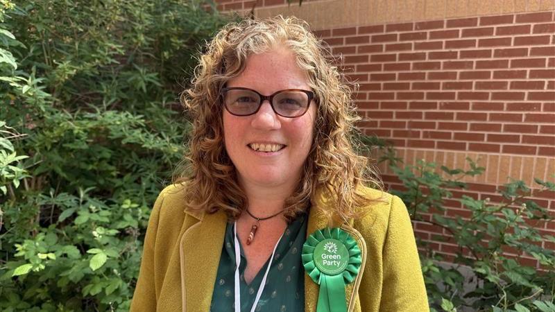 A woman with shoulder-length blonde-brown curly hair and tinted glasses is wearing a dark green blouse under a yellow jacket. She has a green rosette reading "Green Party" on her lapel.