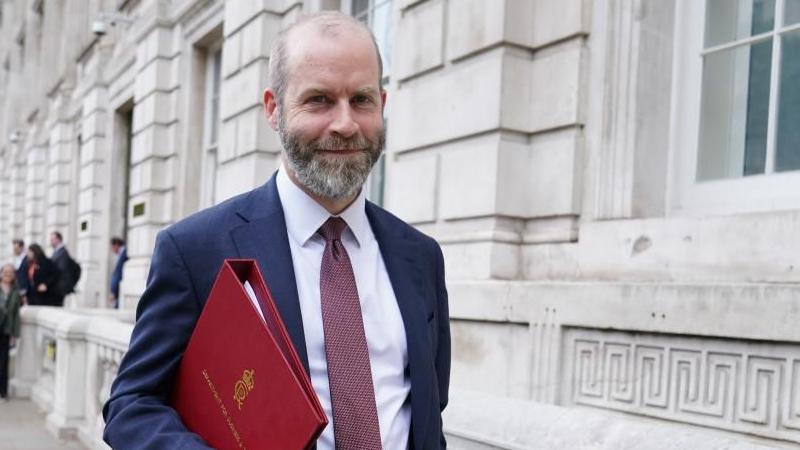 Business Secretary Jonathan Reynolds - he has a greying beard and is wearing a dark blue suit with white shirt and red tie. He is carrying a red ministerial folder