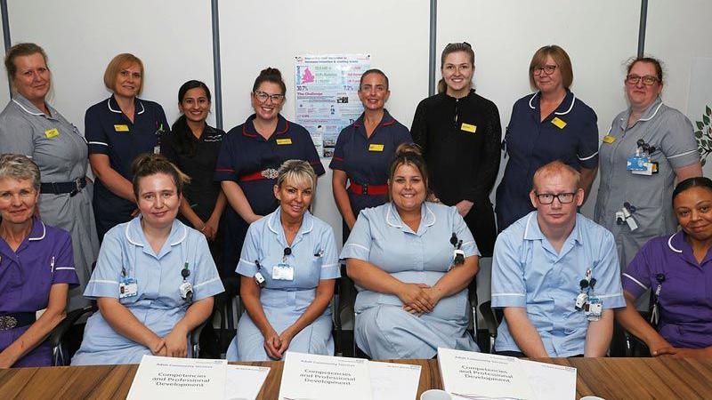 Fourteen members of staff in a range of grey, blue and purple nursing uniforms, one row sitting in chairs behind a desk with papers on, and another standing behind them.  