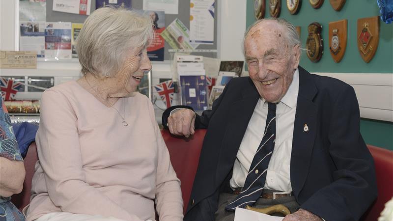 An elderly man is sitting on a chair wearing a suit and tie. He is smiling and holding a walking stick and piece of paper in his hand. Next to him is a woman in a pink jumper smiling at him.